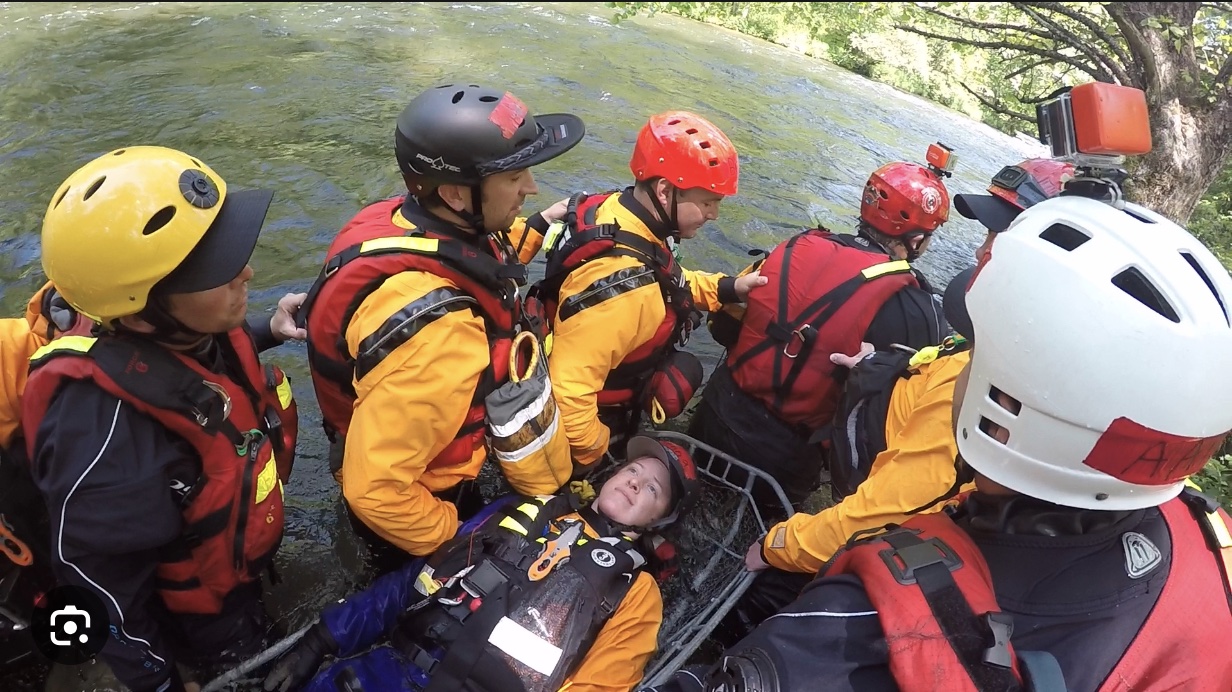 This is a photo from the South Orange Rescue Squad showing six people wearing yellow or black dry suits, red lifevests and red, yellow or white helmets wade through a shallow river carrying a person in a water rescue basket on a training mission for swiftwater rescues.