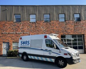 A photo of a white ambulance with dark blue lettering that reads S-O-R-S South Orange Rescue Quad in front of a two-story brick building against a Carolina blue sky, taken by Gabi Battaglini.