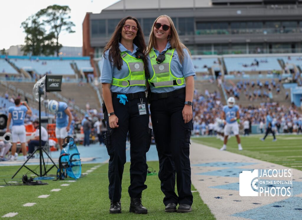 This photograph by SORS board member Jacques Morin shows two South Orange Rescue Squad volunteers standing on the sidelines of a Carolina football game in Kenan Stadium in Chapel Hill, N.C. They are wearing light blue SORS logo shirts, radio sets, dark blue pants and black shoes, sunglasses and high-visibility safety vests. The green grass field with white yard markers and the signature Carolina argyle fills the lower half of the photo. In the bottom right corner is a watermark that reads: Jacques on the sidelines photos with a camera icon. In the immediate background, coaches, trainers and are on the field and the sidelines, along with a parabolic mic and video stand on the left. Farther back, the Carolina blue stands are partially full with fans, who are backed by a Carolina Blue sky and an administrative building. These volunteers are part of the team that staffs every Carolina home football game, providing minor medical assistance and emergency response.