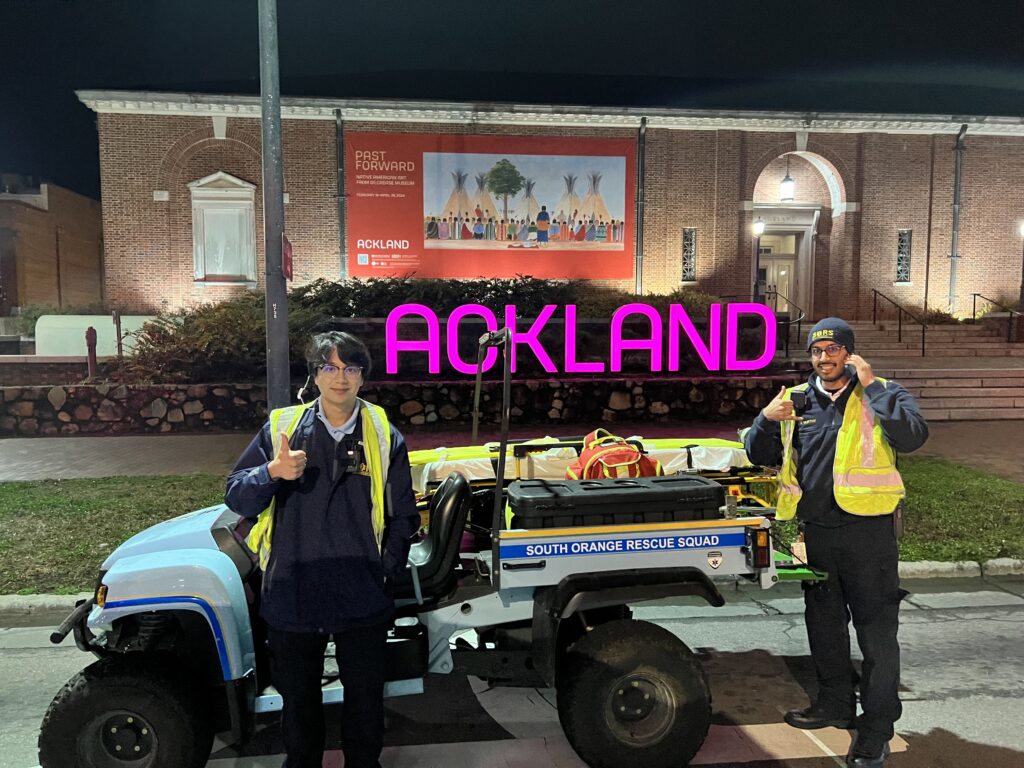 Two young men stand by a gator ATV tricked out as an emergency vehicle with first aid supplies and a stretcher. The gator is light blue with SOUTH ORANGE RESCUE SQUAD written along the side of the bed. The men are wearing dark blue jackets and pants, dark shoes, radio sets and high-visibility vests. They’re smiling and giving the thumbs on. The man on the right is talking on a phone. They are in the street in front of the Ackland Art Museum in downtown Chapel Hill. A bright pink backlit sign reading ACKLAND is on a stone wall that runs along the middle of the photo before stopping at the granite entry steps to the brick building. It’s nighttime, so the sign is vibrant and the building’s facade is uplit against the dark sky, highlighting a false window and tympanum on the right of the building, two side lights and the arched entryway with a hanging light fixture and glass door. A red banner on the facade behind the ACKLAND sign promotes the current exhibition.