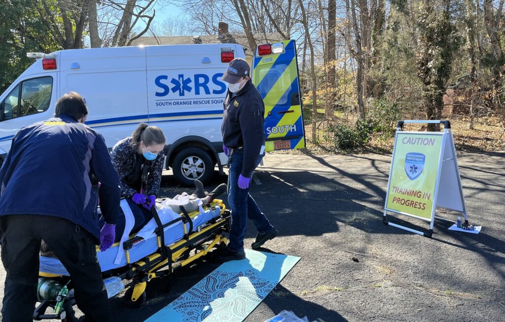 Two emergency medical technicians lean over a person on a stretcher while a third responder looks on in this photo by SORS member Leyla Ozelkan. The standing EMT and the responder in the foreground are wearing dark blue head-to-toe, purple medical gloves and surgical masks. The standing EMT is also wearing a SORS logo hat. The third responder is a woman in a dark patterned sweatshirt with her dark hair in a ponytail. She’s also wearing a mask as she tightens straps about the patient, who is wearing a dark long-sleeved top, tan pants and dark shoes. They are lying on a mattress pad covered in Carolina blue and are partially covered by a white drape. The stretcher is safety yellow. On the ground is a yoga mat and a box of medical supplies. In the background is a SORS ambulance, white with a blue racing stripe and the SORS logo. The back door is open, adorned with blue and safety yellow stripes. The emergency lights are also visible along the top of the rig. In the far background is the roof of a house with a vent and chimney. Bare trees indicate the season is winter. On the right side of the image are some hardwood trees, a small holly and some green ivy. A white sandwich board bears a safety yellow sign reading “CAUTION TRAINING IN PROGRESS” in red letters with the SORS shield logo. The sky is a lovely light blue.