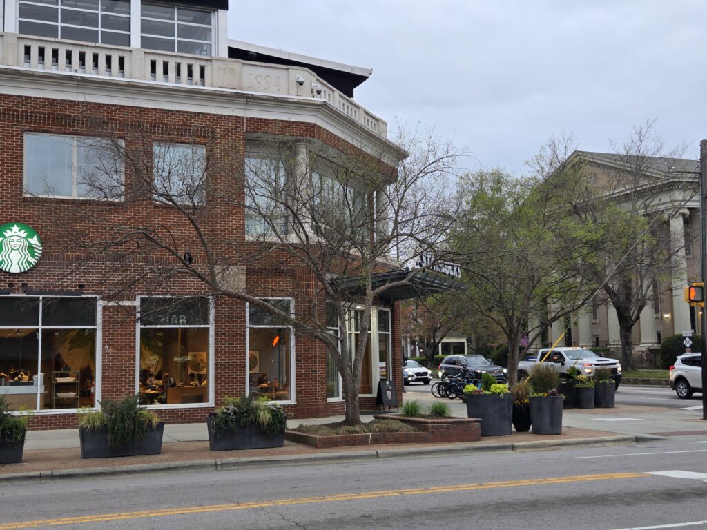 A two-story tall Chinese elm (Ulmus parvifolia) grows out of a brick planter box surrounded by concrete and brick sidewalk in front of a busy coffee house on the southeast corner of Columbia and Franklin Streets in downtown Chapel Hill, N.C. The building has big windows and decorative concrete “railing” along the top. A green and white corporate logo of a woman and a starry crown is on the left. To the right are several cars waiting at a stoplight and the façade of a large church across the street. Several other unidentified trees are visible. The sky is light gray. This photograph was taken by Geoffrey Neal.
