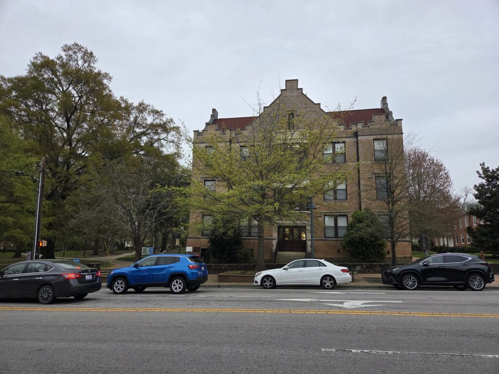 A tan-colored building with a red roof and dark brown window trim and a brown double door sits behind three oak trees, a white oak, a laurel oak and (probably) a pin oak. All three are in various stages of leaf out. The sky is gray and cars are parked on Franklin Street in front of the trees. The photo is by Geoffrey Neal.