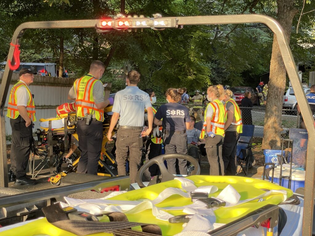 Against a background of green trees and firefighters leaning against a car in the distance, six first responders stand next to a stretcher in front of a couple of seated coworkers discussing a rescue situation. EMS workers are wearing blue shirts and some are also in safety vests. A stretcher is on their left. In the foreground is an all-terrain vehicle with a stretcher. Its illuminated rollbar frames this photo by SORS Chief Matthew Mauzy.