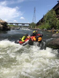 A river rages as two SORS rescuers in a red and black raft assist a kayaker who’s wearing an orange drysuit, black lifevest and helmet. The kayaker is on some river rocks. The rescuers are tethered to a line stretched from one shore to another and are wearing safety yellow waterproof jackets, life vests and helmets. A vehicle bridge and power tower are in the background, A railroad bridge is on the right shore with some trees and rocky shore beneath tit. Another rescuer and a blue kayak are visible on the shore. On the left is a rocky outcrop. Two series of rapids are upriver. This photo was taken by SORS Chief Matthew Mauzy.