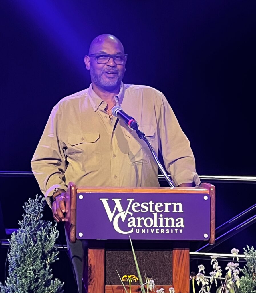 This is a photo of a smiling J. Drew Lanham standing behind the podium during his keynote address at the 2025 Cullowhee Native Plant Conference in July. He wears glasses and a tan button-down shirt with big pockets. A microphone extends in front of him. A placard on the podium reads Western Carolina University. Some native plants are seen near the bottom of the photo. The background is black with some staging in the frame. Photo by Margot Lester.