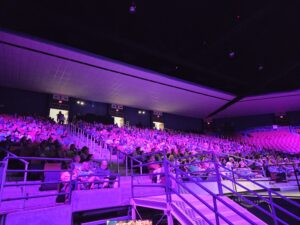 Hundreds of people sitting in the stands of WCU’s Ramsey Center waiting for a plenary session to start at the Cullowhee Native Plant Conference. The crowd is bathed in a purple-ish light because purple is the university’s color. Photo by Geoffrey Neal.