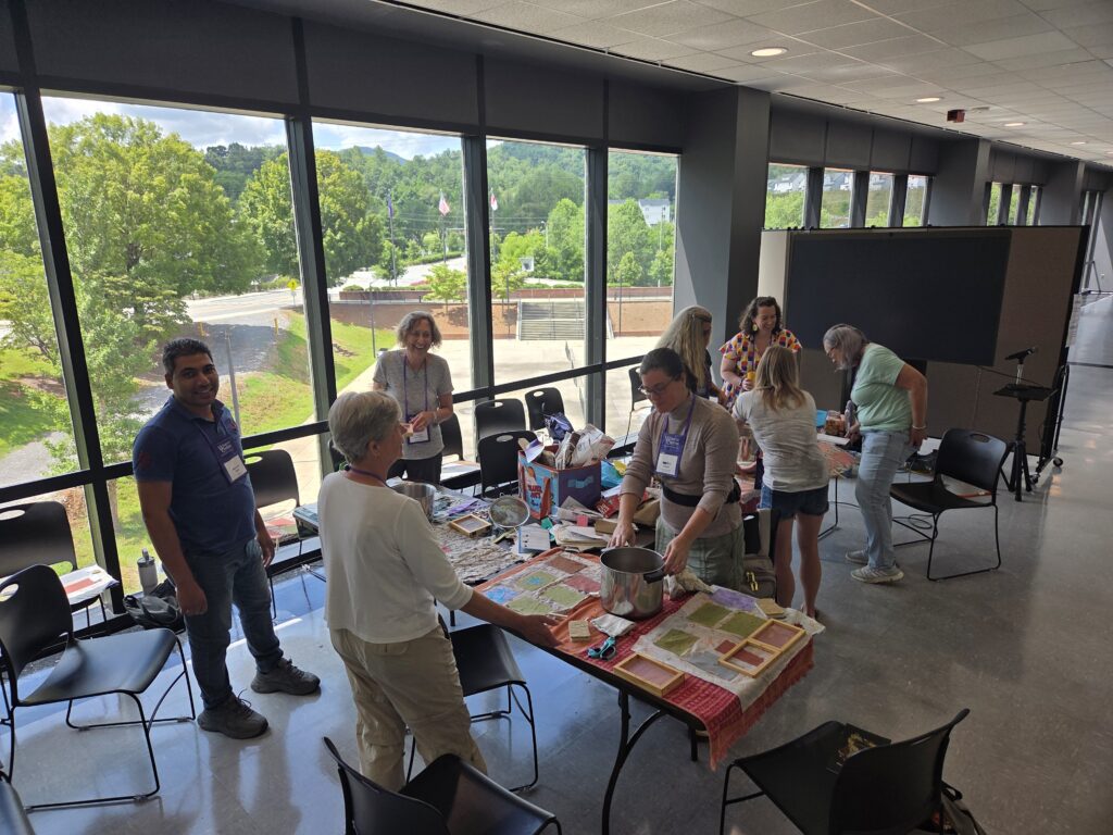 Elizabeth Evans leads a group of people making plantable seed paper at several tables in front of large floor-to-ceiling windows onto a road, trees, a grassy hill and a concrete pickleball court. A brick retaining wall and concrete steps are to the right. The tables are covered in bright cloth with colorful paper and tools. Photo by Geoffrey Neal.