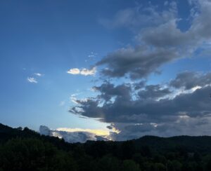 Clouds in shades of gray dominate most of the left side of this photo by Margot Lester. The bottom of the photo is a forested mountaintop in Cullowhee, N.C. Just above the treeline, the last rays of the summer sun shine yellow. To the left, the sky is blue.