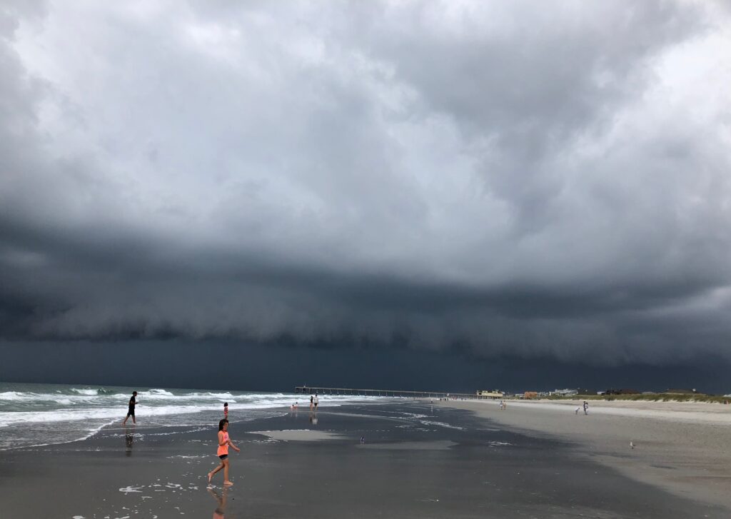 A gigantic dark gray cloud, surrounded by lighter grey ones, stretches from over the Atlantic Ocean on the left past a pier, beach houses and a hotel on the right, menacing waders who are running onshore to get out of the oncoming storm. This photo was taken at Wrightsville Beach, N.C., by Margot Lester.