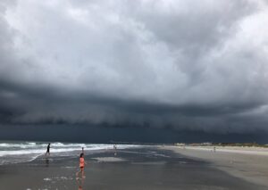 A gigantic dark gray cloud, surrounded by lighter grey ones, stretches from over the Atlantic Ocean on the left past a pier, beach houses and a hotel on the right, menacing waders who are running onshore to get out of the oncoming storm. This photo was taken at Wrightsville Beach, N.C., by Margot Lester.