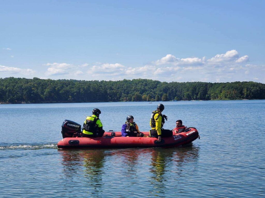 A red rubber zodiac-style raft rides along the surface of a lake on a water-rescue training mission for the South Orange Rescue Squad’s Technical Rescue Team. Four people are aboard, three rescuers wearing lifevests and helmets and an “actor” who needed rescuing. This photo was taken by SORS member Sahil Sethi.
