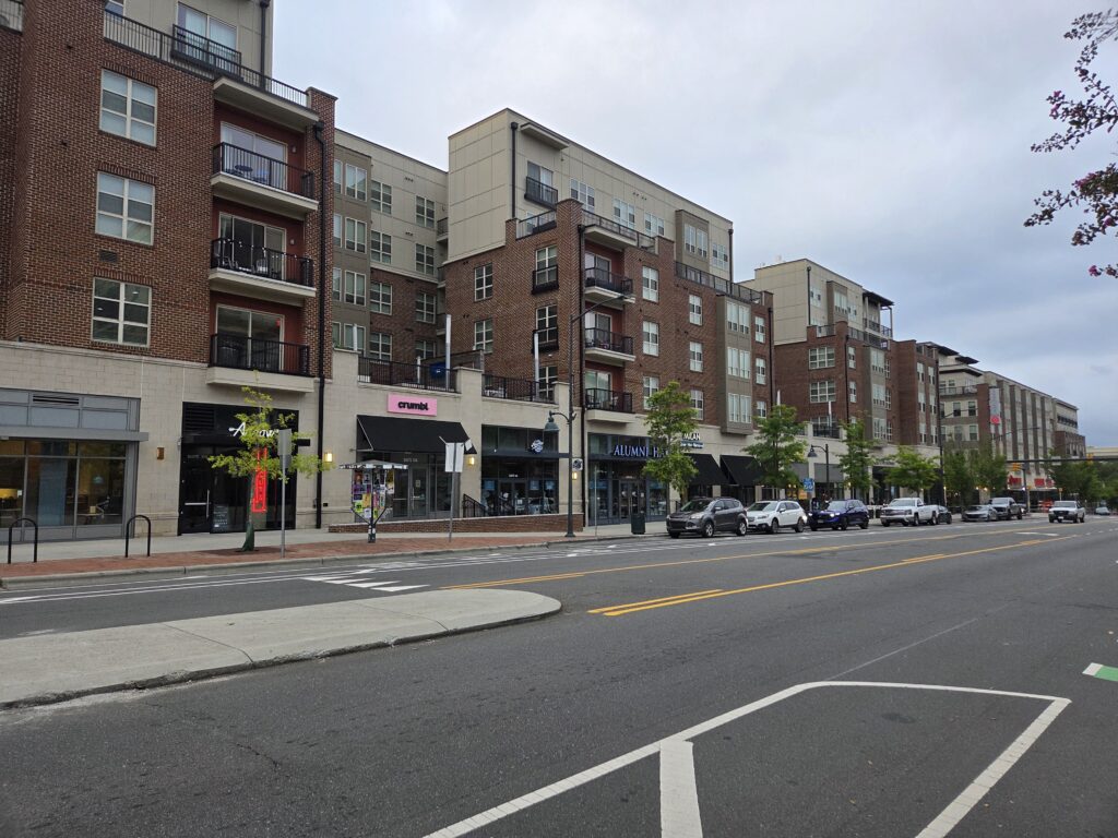A large brick and masonry building rises over West Franklin Street against a blue-gray clouded sky. Between the structures and the car-lined street is a narrow line of youngish trees with green leaves. Photo by Geoffrey Neal.