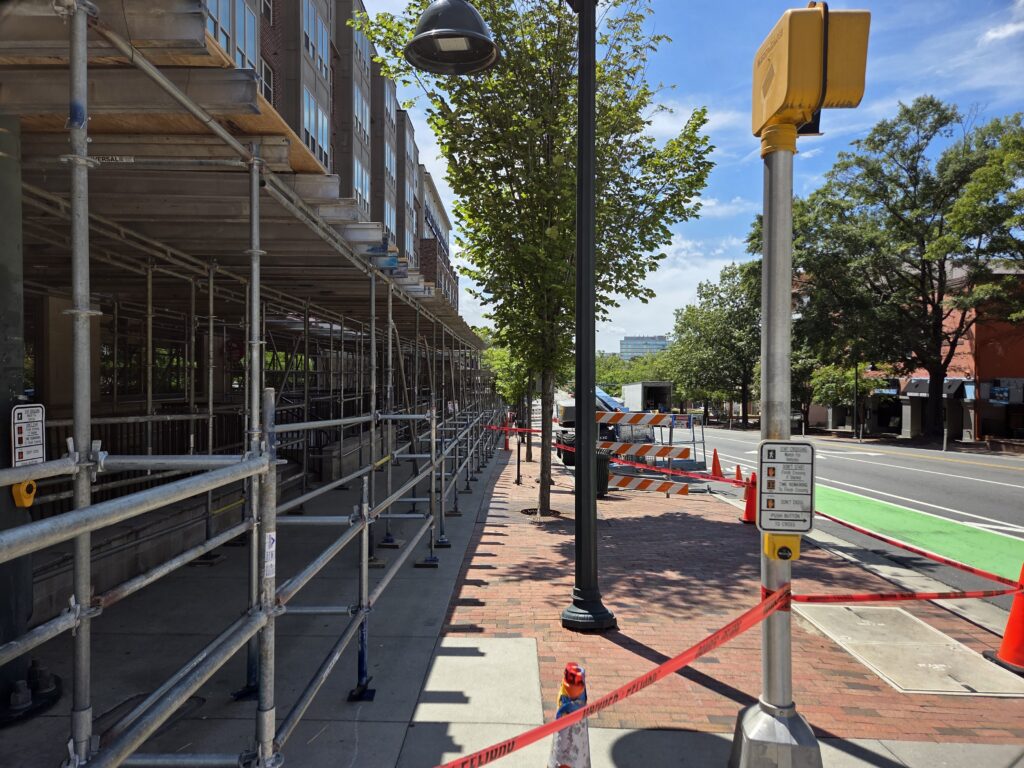 Construction tape, safety cones, barriers and scaffolding crowd a line of street trees in Downtown Chapel Hill. In the foreground is a crossing signal and a lamppost on a concrete and brick sidewalk. To the right are Franklin Street’s bike and vehicle lanes. Older trees and another brick building stretch along the right side of the photo. A large building looms in the distance against a mostly blue sky. 