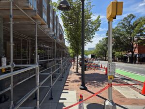 Construction tape, safety cones, barriers and scaffolding crowd a line of street trees in Downtown Chapel Hill. In the foreground is a crossing signal and a lamppost on a concrete and brick sidewalk. To the right are Franklin Street’s bike and vehicle lanes. Older trees and another brick building stretch along the right side of the photo. A large building looms in the distance against a mostly blue sky.