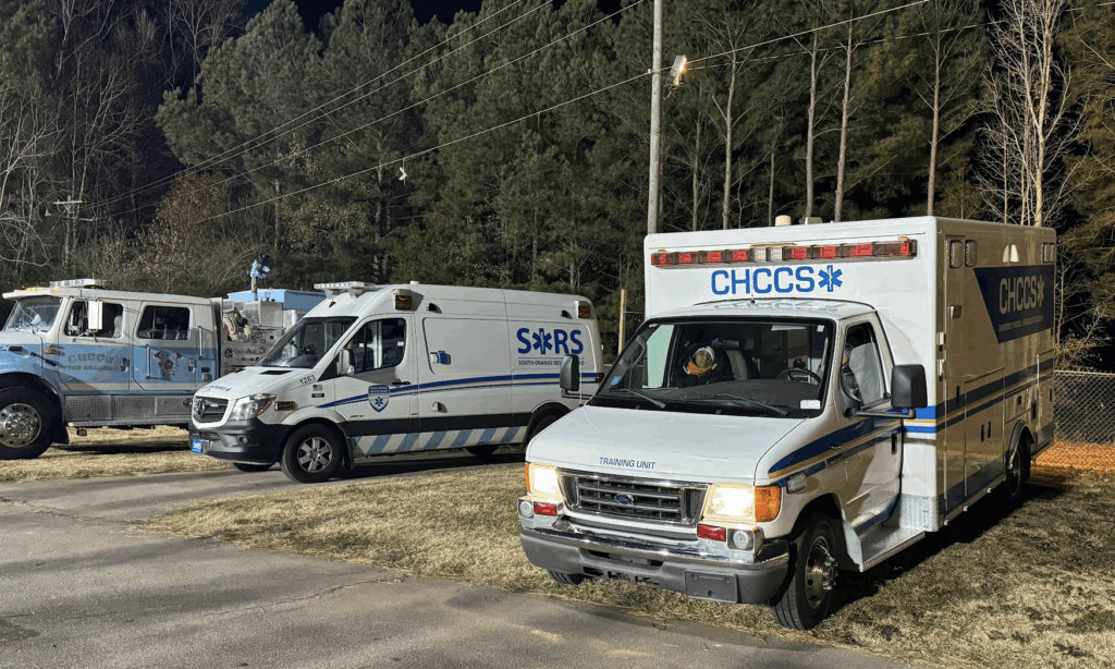 Two emergency response vehicles (a Carolina blue fire truck, far left, and an white boxy ambulance, far right) bearing CHCCS logos, signifying they’re part of the school district’s career and technical education program. An active SORS ambulance is flanked by the two donated vehicles in this nighttime photograph by Gabi Battaglini.