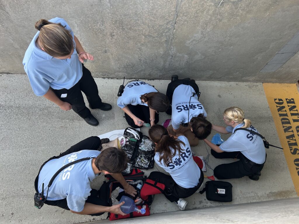 Four EMTs surround a CPR mannequin in a concrete passageway during a simulation drill at Kenan Stadium on the UNC-Chapel Hill campus. To the right, “NO STANDING” is painted in black on a yellow background. Medical supplies are spread out in the foreground. To the left, a SORS volunteer leans over a first aid kit while another stands, providing guidance. SORS Member Reagan Fernandez took this photo.