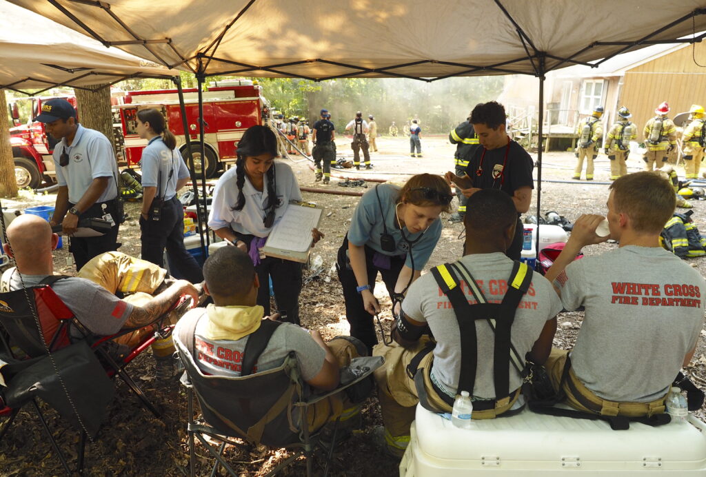 Firefighters stand in front of a burning building while, in the foreground, EMTs from South Orange Rescue Squad check vitals signs and keep firefighters hydrated in a “rehab” tent. Photo by Fred Stipe.