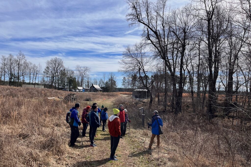 This photo by Margot Lester shows hikers listening to Arborist Geoffrey Neal talking about how to identify trees in the wintertime. The people are standing between a field of brown grasses and a forest of trees. The February sky is a bright blue.