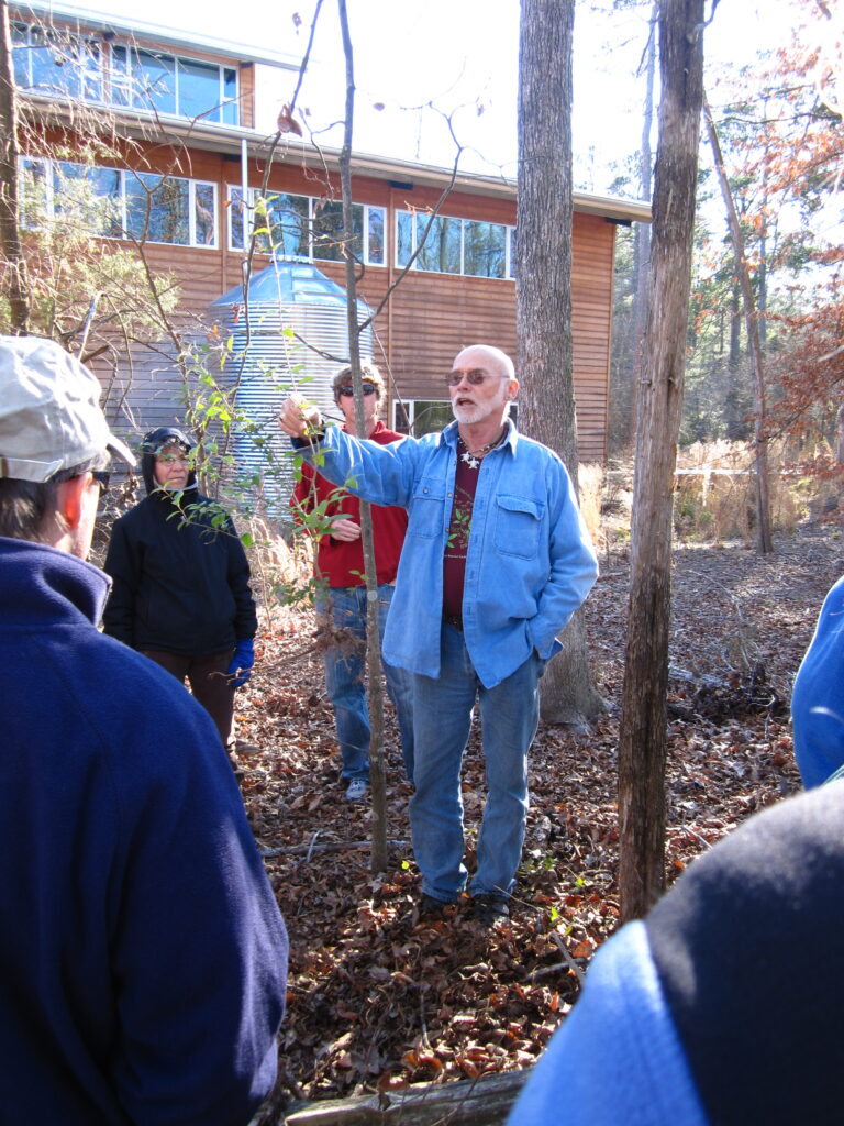 An august gentleman, botanist Ken Moore (in blue jeans, hiking shoes, a light blue chamois shirt and a burgundy t-shirt) holds up a plant for a group of onlookers to study. They are standing in a winter wooded area beside a two-story clapboard building and a large cistern at the North Carolina Botanical Garden in Chapel Hill, N.C.. Photo by Geoffrey Neal.