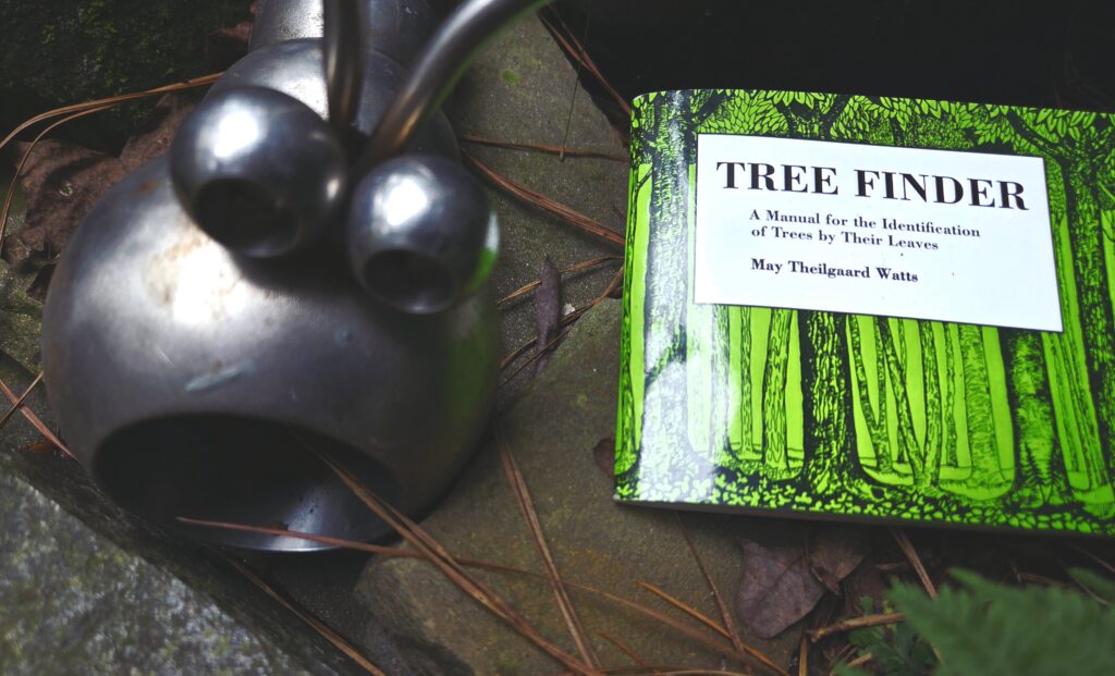 A whimsical insect sculpture, mouth wide open and antennae pointing up, sits next to a book with a shiny green cover illustrated with various tree trunks. Text in a white box announces the title: “Tree Finder. A manual for the identification of Trees by Their Leaves. Amy Theilgaard Watts.” Pinestraw is scattered around the background. Photo by Geoffrey Neal.