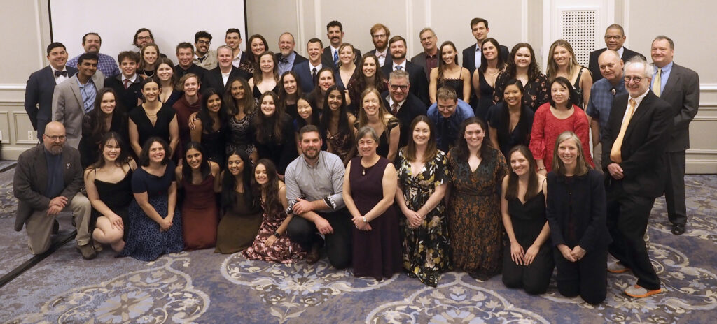 A large group of people in party attire stand or kneel shoulder to shoulder in a hotel ballroom.