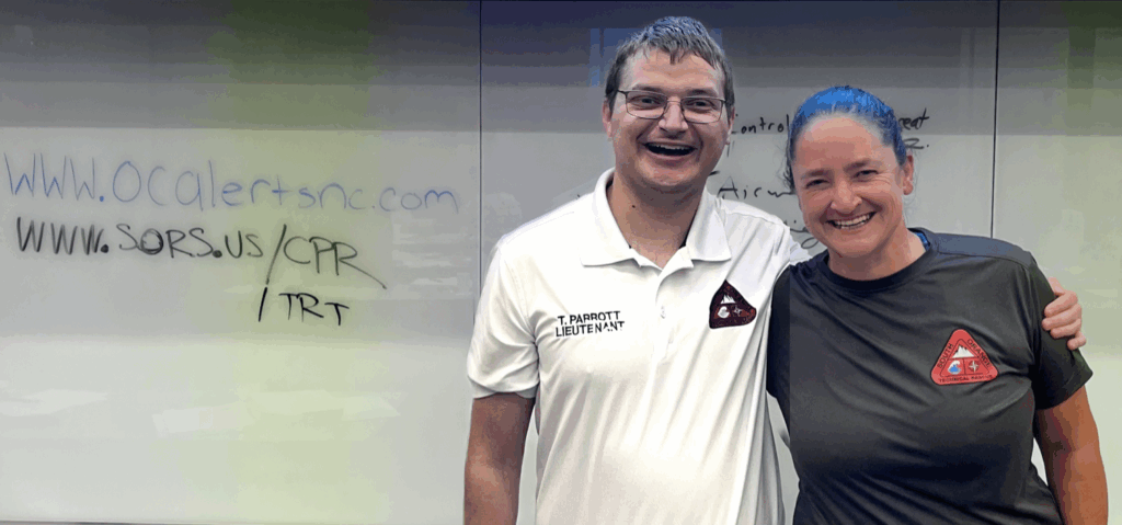 Two first responders (male, left; female, right) in casual shirts stand in front of a white board smiling. One the board is written: www.orangencalerts.com and www.sors.us/CPR and /TRT. This photograph is by Margot Lester.