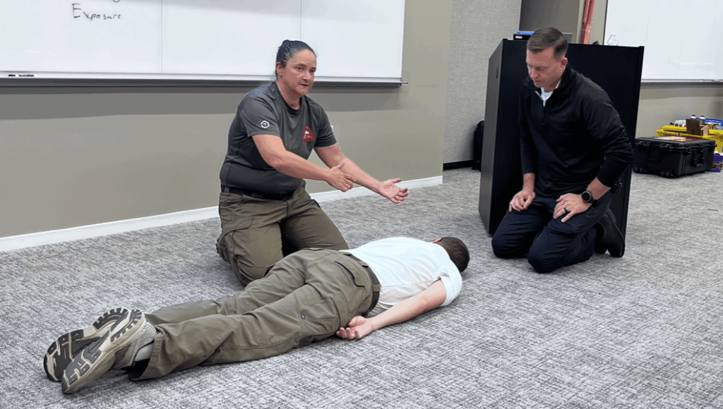 This photograph by Margot Lester show two first responders (female, left; male, right) kneel around another male responder lying face down in a conference room, pretending to be a disaster victim. Behind them is a white board and a lecturn. in casual shirts stand in front of a white board smiling. One the board is written: www.orangencalerts.com and www.sors.us/CPR and /TRT. This photograph is by Margot Lester.