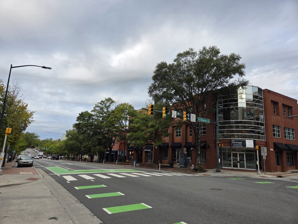 A wide street cuts through this photo of West Franklin Street in downtown Chapel Hill. Parked cars line the left side of the street in front of a row of trees, their leaves starting to change color. A row of large willow oaks lines the right side in front of the three-story brick building with a large, arced window and a for lease sign. A blanket of pale gray clouds covers the light blue sky. Geoffrey Neal took this photo.