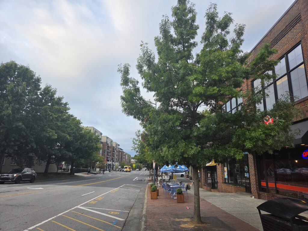 In this photograph of downtown Chapel Hill, N.C., by Geoffrey Neal, a brick-and-glass building facade dominates the right side, punctuated by a few bright blue-and-white umbrellas covering outside seating. Several large trees and a mostly empty street are to the left. Beyond rises a city block of more brick buildings with a school bus making its way. Smaller trees are also visible. The sky is pale blue with some puffy white-ish gray clouds.