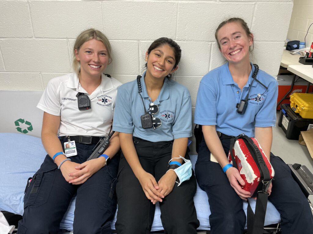 Three women sit smiling on a Carolina blue cushion in front of a white cinderblock wall. The woman on the right has blonde hair and is wearing a white short-sleeved shirt and dark pants. She’s holding a walkie-talkie. The middle woman is wearing a light blue shirt and dark pants. She has dark brown hair and has a face mask around her wrist. The woman on the left has light brown hair and is also dressed in a light blue shirt and dark pants. She’s holding a portable automatic external defibrillator unit in a red and white pouch. They are SORS volunteer EMTs at a University of North Carolina sporting event. 