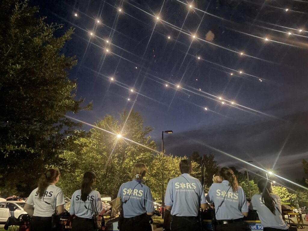 This photo features six SORS volunteer EMTs standing in a parking lot staring up a fireworks ablaze in the dark blue sky. A bank of purple-ish-gray clouds is in the bottom right corner. Green trees run left to right through the middle of the photo. Two streetlights also flare in the midground. 