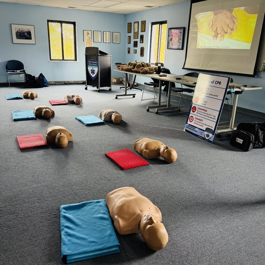 This photo shows two rows of CPR mannequins, which are realistic head-and-torso models with sensors inside that help us teach people life-saving CPR. Blue and red mats lie beside them to cushion trainees’ knees. An instructional sign leans against a conference table. Nearby are more supplies for the class. On a pull-down screen, there’s an image of hands with fingers entwined, ready to give CPR to a mannequin in a yellow shirt. This is the training room at the SORS station in Carrboro, N.C. 