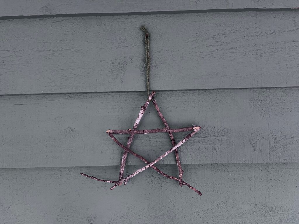 Five sticks are glued together to form a star hung by green landscape twine on grayish-green clapboards. These were made and photographed by Margot Lester.