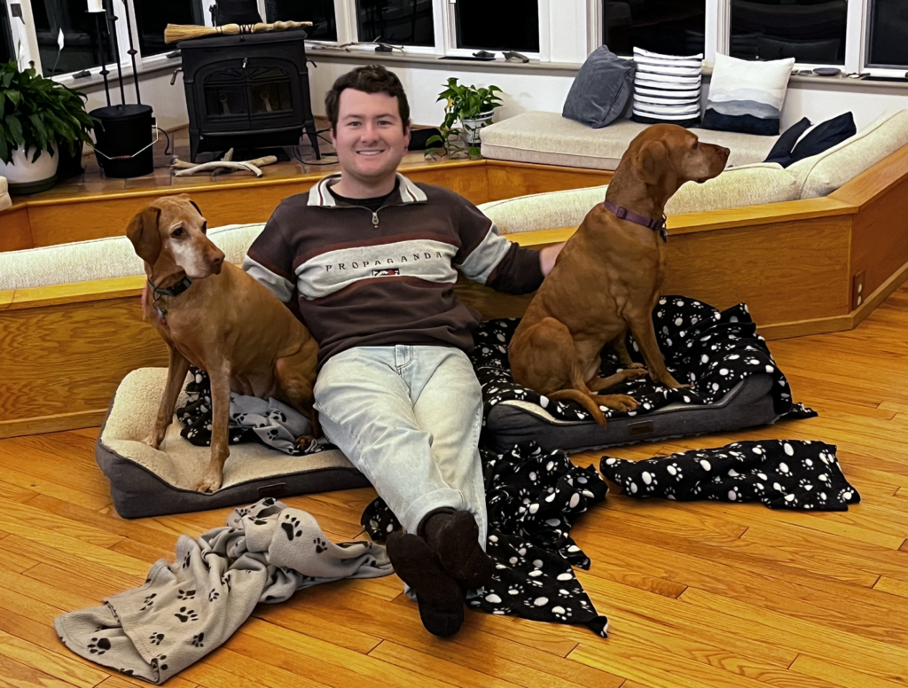 This is a photo by SORS volunteer Margot Lester of a happy young man in his mid-20s (with dark hair, wearing khakis and a brown sweatshirt) sitting between two rust-colored older Viszla dogs in their comfy beds.