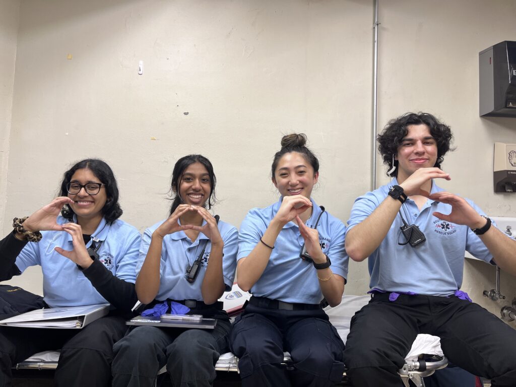 Four young EMTs sit on a stretcher against a plain white wall. They wear dark pants and light blue shirts. Nitrile gloves are visible, as is their comms gear. Their hands are shaping the letters S, O, R, S. Photo by SORS Member Soorya Vasan