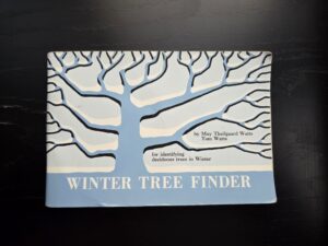 A small book sits on a black counter. Its cover, in shades of blue, is an illustration of a large tree in winter. Its light blue trunk rises wide from the ground against a paler blue background. White snow caps its branches and black lines represent shadows. The title, Winter Tree Finder, is written in black all-caps serif type along the bottom. The words “by Mary Theilgaard Watts and Tom Watts” appear on the right between a forked branch in the same font. Below that, between the bottom branch and the snow-covered ground, is black type that reads: “for identifying deciduous trees in Winter”.