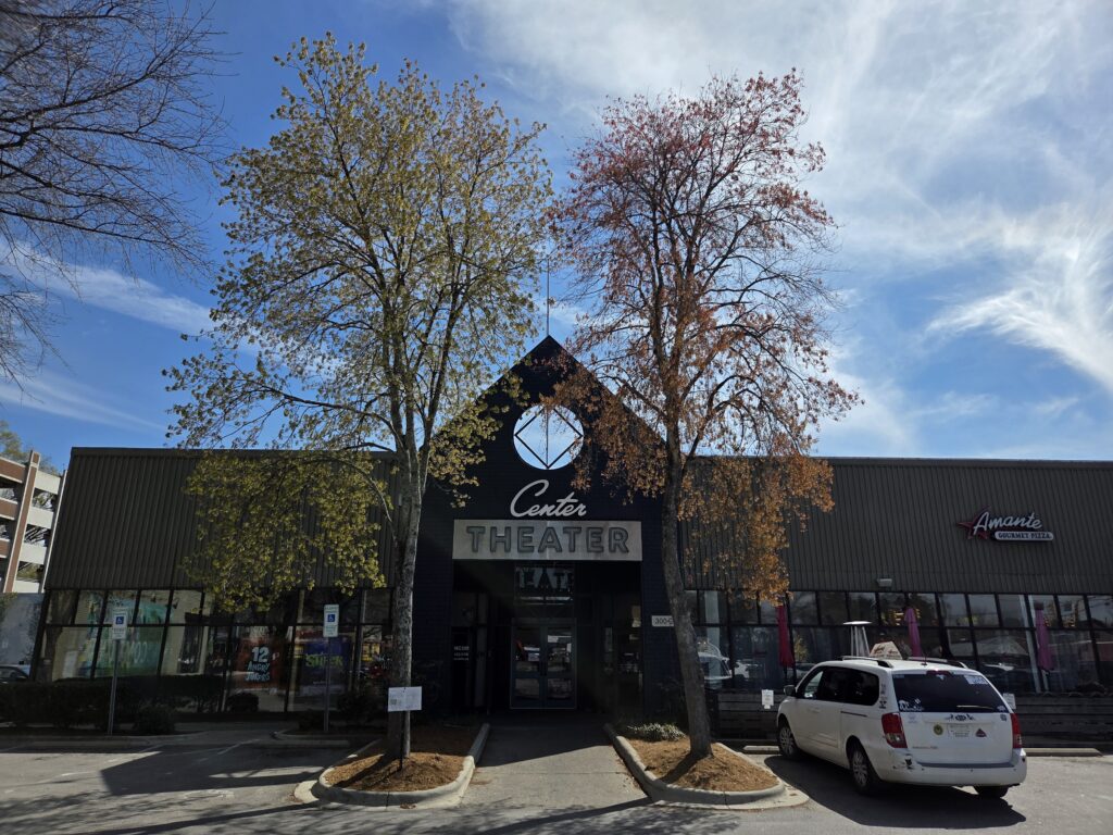 Two healthy maple trees flank the entrance to the Carrboro, N.C.’s Center Theater, backed by a blue sky and wispy white clouds. The trees are showing off their spring colors: samaras or fruits that are excellent flyers, spreading maple seeds with every good wind. The building’s roofline features a large triangle with a “porthole” centered over a sign reading “Center Theatre”. A delivery van is also in the foreground.