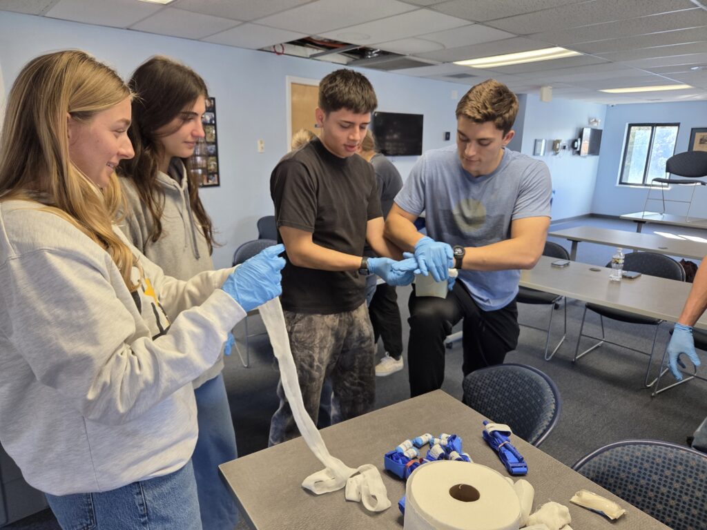 Two young women (left) look on as two young men practice putting gauze pads and pressure on a simulated injury. Everyone is wearing blue nitrile gloves. One of the women is holding a partially unrolled gauze strip. On the table in front of them are various supplies used in the class. 