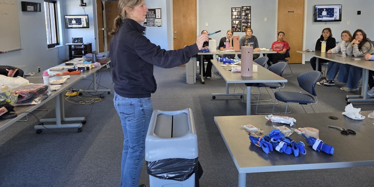 A woman stands in a training room between tables filled with tourniquets, gauze and other materials for a Stop the Bleed training session.