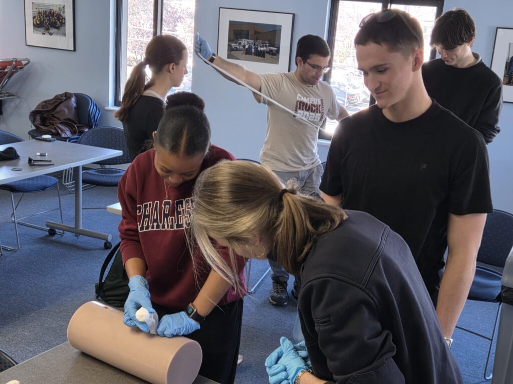 Participants in a recent SORS Stop the Bleed training practice treating different kinds of injuries. In the foreground, a young woman inserts a roll of gauze into a round flesh-colored cylinder that simulates a human leg as the instructor (center) and other participant (right) look on. Another team works in the background. Two participants in dark shirts flank a man in a tan shirt who’s stretching cotton between his outstretched arms. 