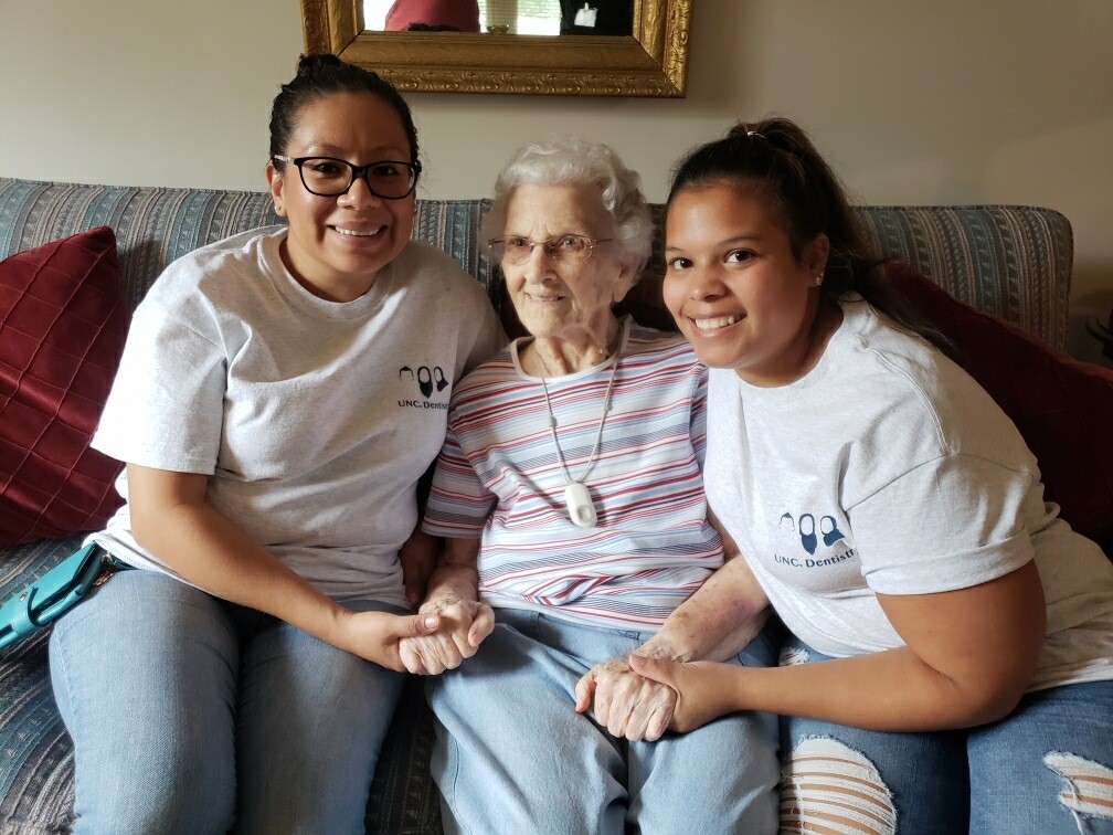 Two young women side on either side of an older woman in her home.