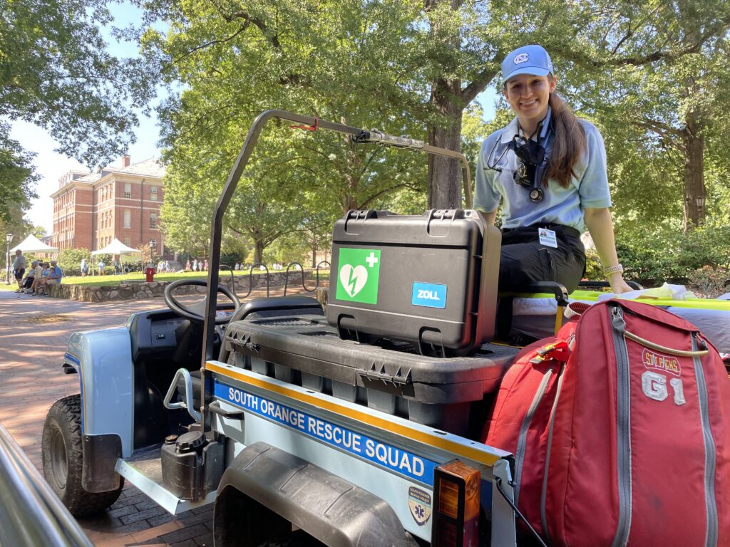 A smiling young EMT sits on top of an all-terrain vehicle full of rescue equipment on a summer evening. The words SOUTH ORANGE RESCUE SQUAD adorn the rig. 