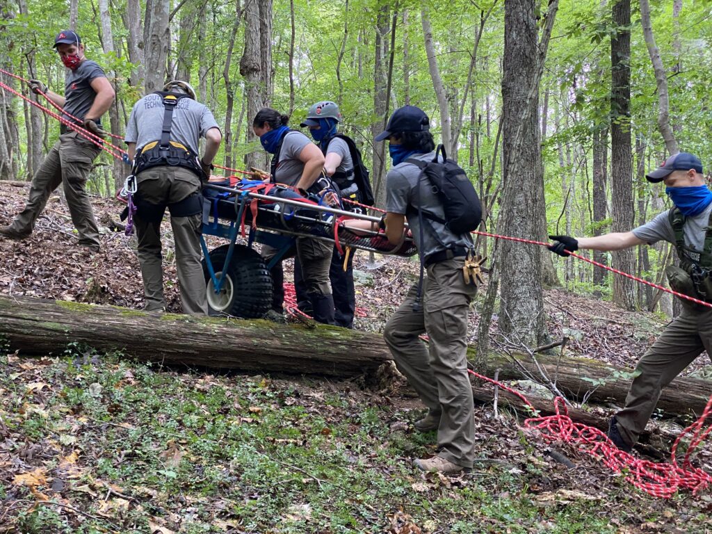 In this photo by SORS Chief Matthew Mauzy, six members of the SORS Technical Rescue Team dressed in rugged outdoor apparel and wearing caps and various sorts of backpacks ferry an injured party in a stretcher up a wooded hillside. They are climbing over a fallen tree. The stretcher is supported by a red rope tethered higher uphill and is rolling on a large tire. The forest floor is a mix of leaf litter, dirt and groundcover plants. Tree trunks and leafed-out limbs make up the background. 