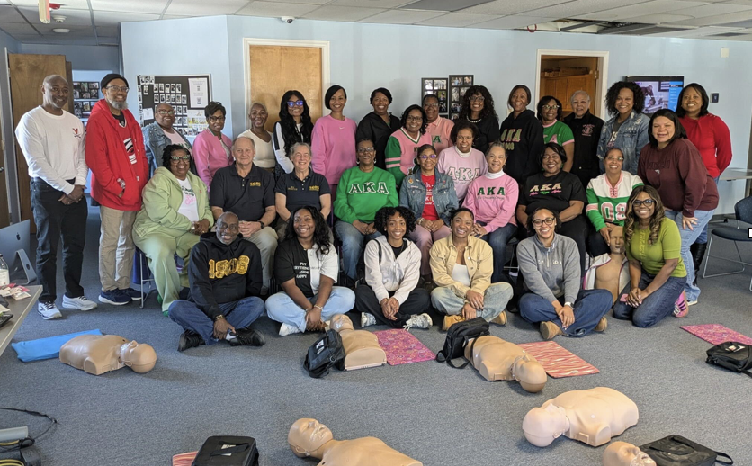 A large group of people, many wearing tops emblazoned with the Greek letters for Alpha Kappa Alpha, stand and sit by CPR manikins in the South Orange (NC) Rescue Squad training room. 