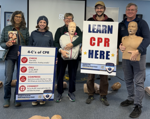 Five adults stand in a conference room holding various CPR training materials including booklets, manikins and signage. 