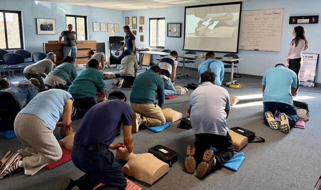 Several adults kneel on a gray carpet performing CPR on mannikins, while instructors stand nearby offering guidance. A video on a drop-down screen and messages on the whiteboard deliver additional instruction in Spanish.