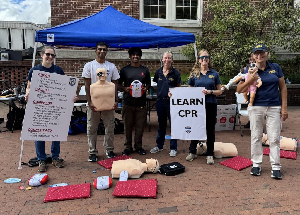 Six smiling members of the SORS CPR team stand in front of a brick building on a brick walkway with signs, and AED and manikins wearing eye patches. 