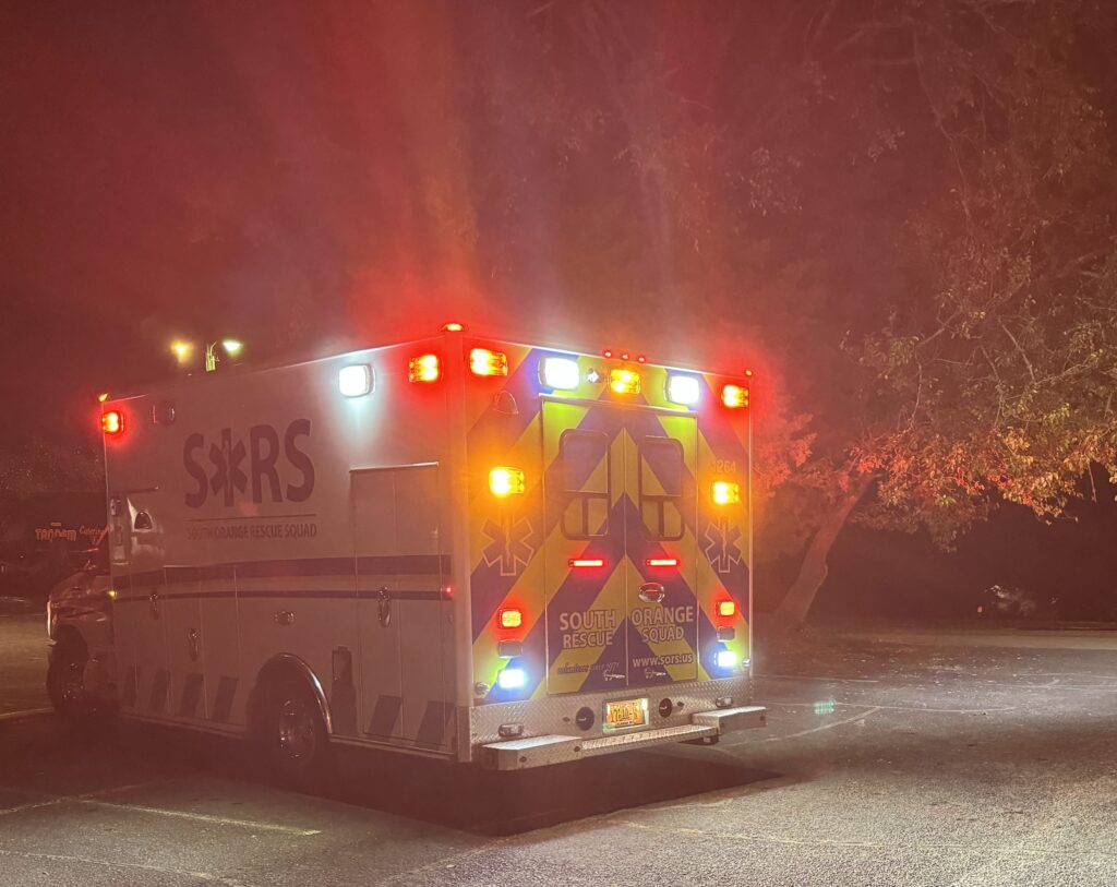 A SORS ambulance, lights aglow, drives off into the night. A large tree is in the background, its understory partially illuminated by the lights. 