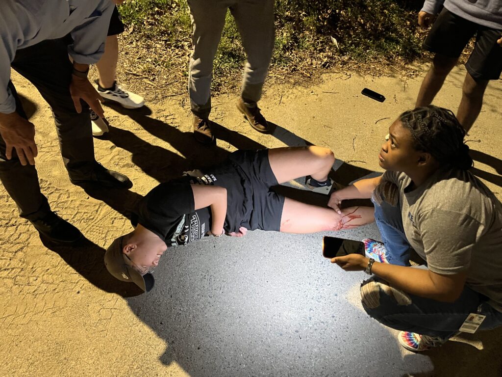 A young man lies injured on a path, his arm pinned under his body. It’s nighttime, so a young woman kneels next to him, using her phone flashlight to illuminate the scene. Four other people stand nearby, one leaning forward, talking to the woman. 